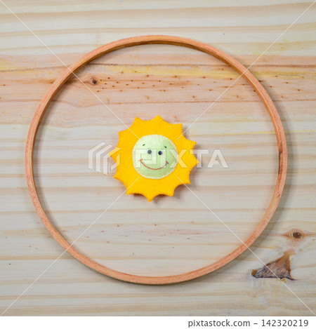 Handmade felt smiling sun decor arranged inside a circular wooden ring on a light wooden background Handmade felt smiling sun decor arranged inside a circular wooden ring on a light wooden background 142320219