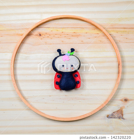 Handmade felt ladybug decor arranged inside a circular wooden ring on a light wooden background Handmade felt ladybug decor arranged inside a circular wooden ring on a light wooden background 142320220