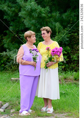 Full length portrait of two senior russian sisters looking at each other and smiling with flower bouquets standing in a garden in Russia Full length portrait of two senior russian sisters looking at each other and smiling with flower bouquets standing in a garden in Russia 142320636