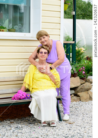 Authentic portrait of two senior russian sisters 80 and 75 years old sitting on a bench near a house porch in a summer garden in Russia 142320637