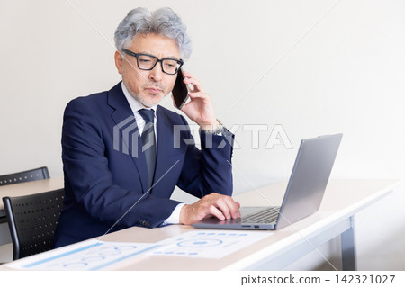 Japanese businessman working on his laptop while talking on his smartphone. Japanese businessman working on his laptop while talking on his smartphone. 142321027