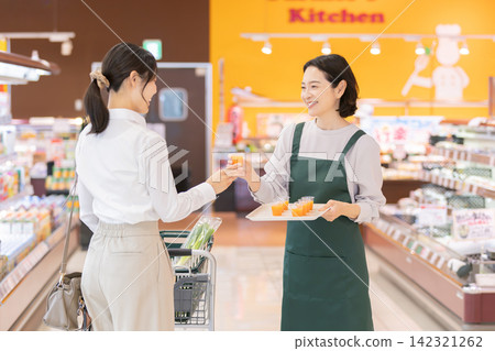 超市裡,一位面帶微笑的女店員向一位女顧客提供免費試飲。 超市裡,一位面帶微笑的女店員向一位女顧客提供免費試飲。 142321262