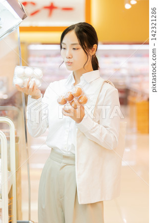 A young woman compares white eggs and brown eggs in the supermarket's egg section to choose one. A young woman compares white eggs and brown eggs in the supermarket's egg section to choose one. 142321286