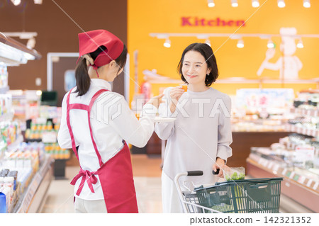 A smiling female staff member offers a free sample drink to a female customer at a supermarket. A smiling female staff member offers a free sample drink to a female customer at a supermarket. 142321352