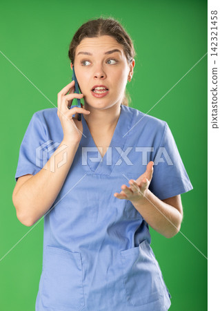 Professional female doctor wearing blue medical clothing convincingly and emotionally talking to patient by smartphone standing against green screen Professional female doctor wearing blue medical clothing convincingly and emotionally talking to patient by smartphone standing against green screen 142321458