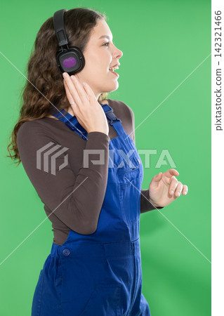 Female worker in a blue overalls happily listens to music on headphones and dances against green background Female worker in a blue overalls happily listens to music on headphones and dances against green background 142321466