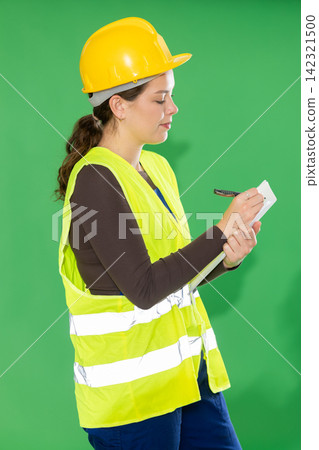 Female worker in reflective vest and hard helmet takes inventory and makes notes on paper on green background Female worker in reflective vest and hard helmet takes inventory and makes notes on paper on green background 142321500