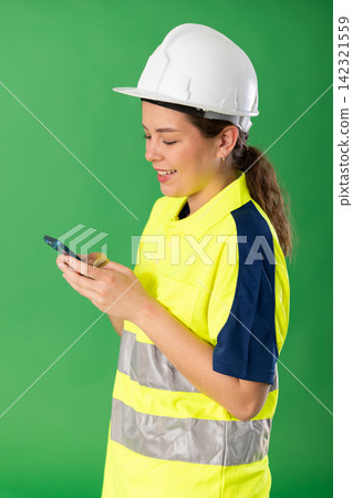 Polite girl worker wearing high visibility polo and hard hat scrolling smartphone standing against green screen Polite girl worker wearing high visibility polo and hard hat scrolling smartphone standing against green screen 142321559
