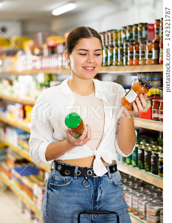 in department of canned vegetables and fruits, young woman chooses small jar of canned carrots in department of canned vegetables and fruits, young woman chooses small jar of canned carrots 142321787