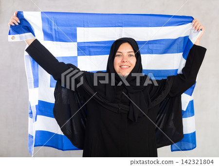 Balanced smiling Muslim woman in traditional black hijab holds flag of Greece Balanced smiling Muslim woman in traditional black hijab holds flag of Greece 142321804