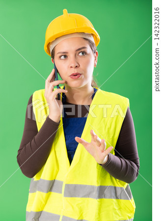 Female worker in reflective vest and hard helmet talking on a mobile phone and giving instructions on green background Female worker in reflective vest and hard helmet talking on a mobile phone and giving instructions on green background 142322016