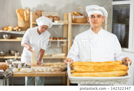 Enthusiastic young baker presenting freshly baked golden baguettes Enthusiastic young baker presenting freshly baked golden baguettes 142322347