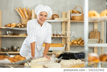 Young baker girl is standing near work table in kitchen, working with dough Young baker girl is standing near work table in kitchen, working with dough 142322442