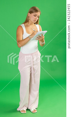 Attentive teenage girl in pink trousers takes notes in notebook on green background. Chroma key compositing Attentive teenage girl in pink trousers takes notes in notebook on green background. Chroma key compositing 142322455