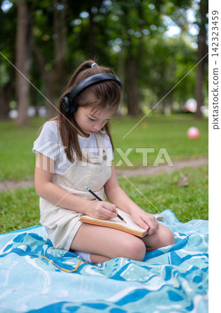 Young girl wearing headphones writing drawing in a notebook while sitting on picnic blanket in park Young girl wearing headphones writing drawing in a notebook while sitting on picnic blanket in park 142323459