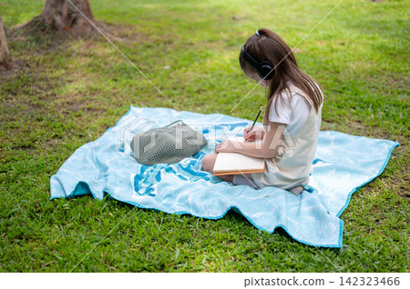 Side view of a young girl wearing headphones writes in a notebook while sitting on a blanket in park Side view of a young girl wearing headphones writes in a notebook while sitting on a blanket in park 142323466