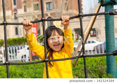 Joyful little girl smiles excitedly while playing inside a rope climbing net at an outdoor playground. Joyful little girl smiles excitedly while playing inside a rope climbing net at an outdoor playground. 142324164
