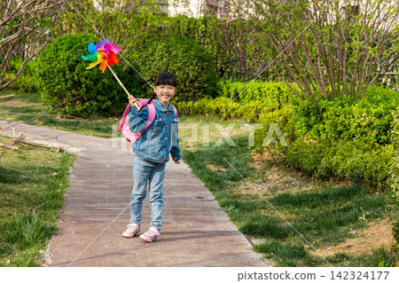 A happy young girl with a pink schoolbag stands on a park path, smiling warmly and holding a colorful pinwheel during a sunny day. A happy young girl with a pink schoolbag stands on a park path, smiling warmly and holding a colorful pinwheel during a sunny day. 142324177