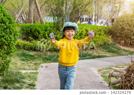 A cheerful little girl wearing a yellow sweatshirt and a sun hat runs toward the camera on a park path, holding small purple flowers. 142324179