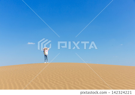 A joyful young girl wearing a white cap and colorful windbreaker walks up a vast sand dune under a clear blue sky. A joyful young girl wearing a white cap and colorful windbreaker walks up a vast sand dune under a clear blue sky. 142324221