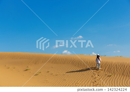 A joyful young girl wearing a white cap and colorful windbreaker walks up a vast sand dune under a clear blue sky. A joyful young girl wearing a white cap and colorful windbreaker walks up a vast sand dune under a clear blue sky. 142324224