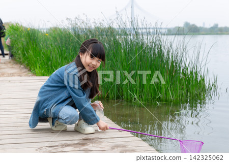 A young girl in a denim jacket was squatting on the wooden boardwalk near the lake, catching fish with a net in her hand. A young girl in a denim jacket was squatting on the wooden boardwalk near the lake, catching fish with a net in her hand. 142325062