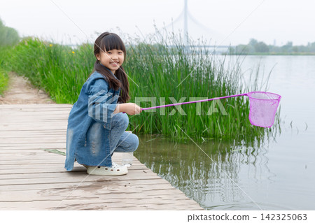 A young girl in a denim jacket was squatting on the wooden boardwalk near the lake, catching fish with a net in her hand. A young girl in a denim jacket was squatting on the wooden boardwalk near the lake, catching fish with a net in her hand. 142325063