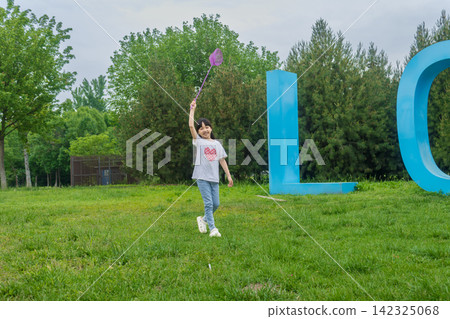 Cheerful little girl holding a purple butterfly net on a spacious green lawn. 142325068