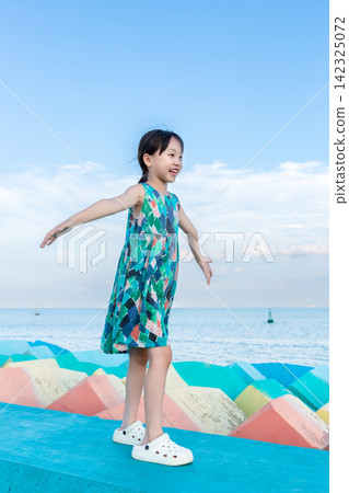Happy little girl wearing a colorful dress standing on a blue seawall with open arms. Happy little girl wearing a colorful dress standing on a blue seawall with open arms. 142325072