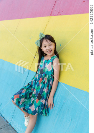 Happy young girl standing in front of a striped pink, yellow, and blue wall. Happy young girl standing in front of a striped pink, yellow, and blue wall. 142325092