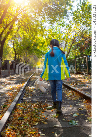 Back view of a child walking down a historic train track carrying a white teddy bear in autumn. Back view of a child walking down a historic train track carrying a white teddy bear in autumn. 142325122