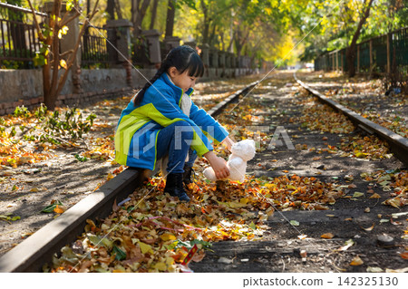 A child squatting down on an autumn railway track to place a white teddy bear among the fallen yellow leaves. A child squatting down on an autumn railway track to place a white teddy bear among the fallen yellow leaves. 142325130