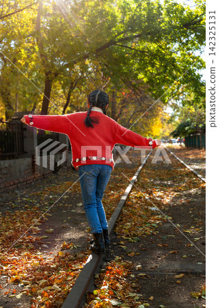 Back view of a child in a red knitted sweater balancing carefully while walking on a single railway rail in autumn. Back view of a child in a red knitted sweater balancing carefully while walking on a single railway rail in autumn. 142325131