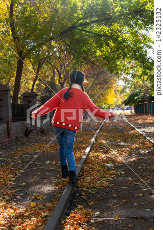 Back view of a child in a red knitted sweater balancing carefully while walking on a single railway rail in autumn. Back view of a child in a red knitted sweater balancing carefully while walking on a single railway rail in autumn. 142325132