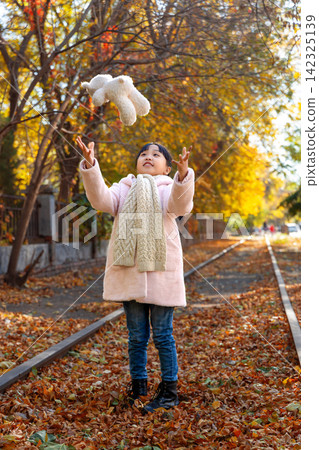 A cheerful young girl throwing a plush white teddy bear up into the air on an autumn railway track. A cheerful young girl throwing a plush white teddy bear up into the air on an autumn railway track. 142325139