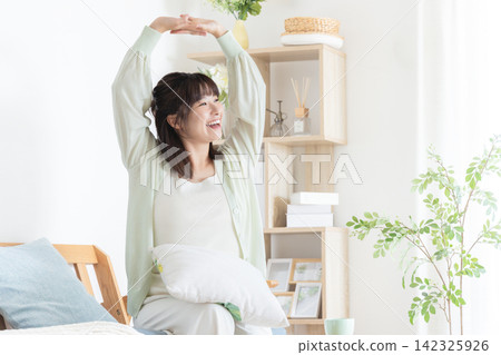 一位微笑的年輕女子在沐浴著晨光的房間裡伸了個懶腰,神清氣爽地醒來。 一位微笑的年輕女子在沐浴著晨光的房間裡伸了個懶腰,神清氣爽地醒來。 142325926