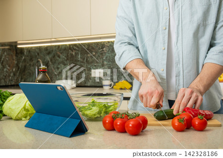 Cooking. Person cuts cucumber and tomatoes in kitchen while using tablet for recipe Cooking. Person cuts cucumber and tomatoes in kitchen while using tablet for recipe 142328186
