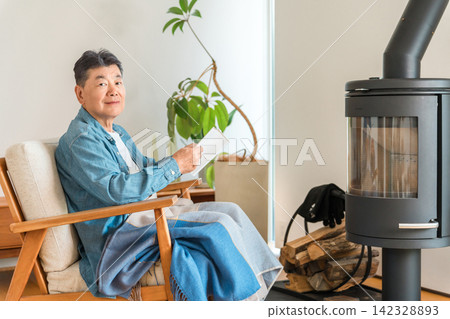 一位老人坐在客廳的燃木爐前的椅子上看書。 一位老人坐在客廳的燃木爐前的椅子上看書。 142328893