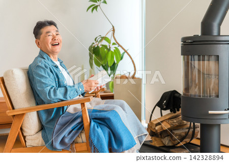 A senior man sits in a chair in front of the wood-burning stove in his living room, reading a book. A senior man sits in a chair in front of the wood-burning stove in his living room, reading a book. 142328894