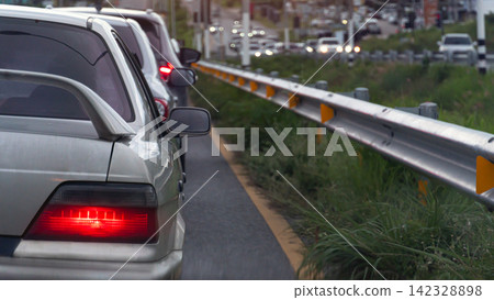 Rear side of silver car stopped in a traffic queue alongside a metal highway guardrail during a daytime commute. With turnon brake light side of guardreal. With green grass for road barrier. Rear side of silver car stopped in a traffic queue alongside a metal highway guardrail during a daytime commute. With turnon brake light side of guardreal. With green grass for road barrier. 142328898
