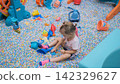Little girl sitting in a sandbox filled with colorful plastic balls playing with a bucket and toy shapes. Indoor creative play concept 142329627