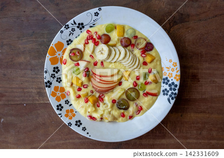 Creamy fruit custard topped with sliced banana apple pomegranate seeds and grapes in plate on wooden table. Delicious homemade sweet dessert served chilled. Top view. Creamy fruit custard topped with sliced banana apple pomegranate seeds and grapes in plate on wooden table. Delicious homemade sweet dessert served chilled. Top view. 142331609