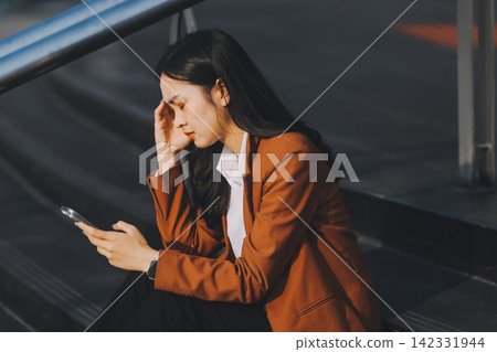 Asian business woman sitting alone on concrete steps, suffering from severe headache, mental stress and deep sadness, contemplating failure after losing her job Asian business woman sitting alone on concrete steps, suffering from severe headache, mental stress and deep sadness, contemplating failure after losing her job 142331944