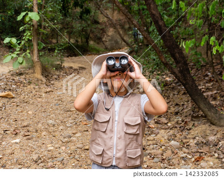 Little Asian girl wearing safari hat and vest looking through binoculars on forest nature trail Little Asian girl wearing safari hat and vest looking through binoculars on forest nature trail 142332085