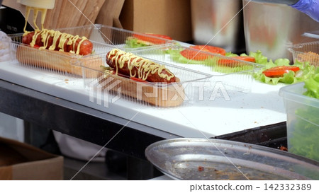 Finished frankfurter topped with mustard and ketchup, melted cheese and garnish sitting on bun ready to serve, golden tones evoke savory aroma, vendor presentation for hungry festival crowd Finished frankfurter topped with mustard and ketchup, melted cheese and garnish sitting on bun ready to serve, golden tones evoke savory aroma, vendor presentation for hungry festival crowd 142332389