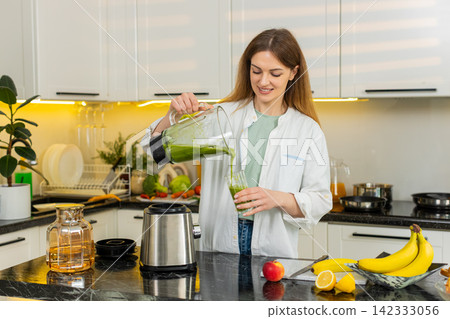 Middle-aged woman pouring fresh green smoothie from blender in home kitchen for detox breakfast diet Middle-aged woman pouring fresh green smoothie from blender in home kitchen for detox breakfast diet 142333056