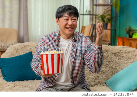 Asian man laughing loudly watching comedy movie eating popcorn, joyful relaxed mood, hearty laughter Asian man laughing loudly watching comedy movie eating popcorn, joyful relaxed mood, hearty laughter 142333074