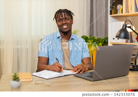 African American young man on home table checks task list and writes notes for laptop work session African American young man on home table checks task list and writes notes for laptop work session 142333078