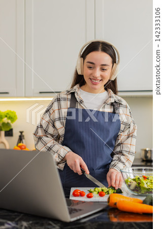 Young woman slicing cucumber and tomatoes for keto salad while viewing recipe on laptop at home meal Young woman slicing cucumber and tomatoes for keto salad while viewing recipe on laptop at home meal 142333106