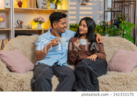 Indian couple man and woman sitting on home sofa drinking morning coffee and enjoying relaxed talk Indian couple man and woman sitting on home sofa drinking morning coffee and enjoying relaxed talk 142333170